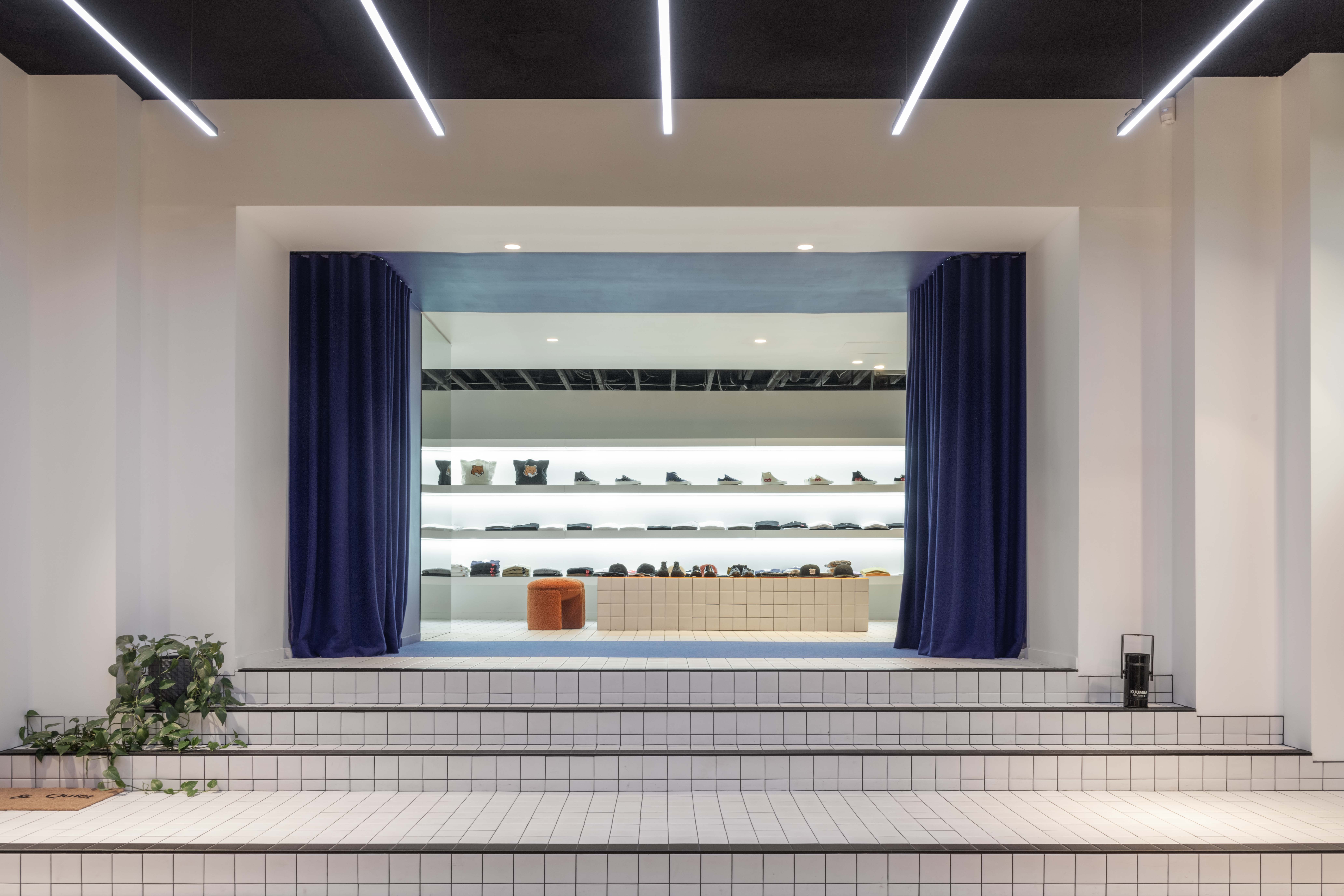 Stage-like tiled staircase inside the 199A® space, framed by deep blue curtains and linear ceiling lights, opening onto a minimalist retail backdrop with sculptural seating and shelving.