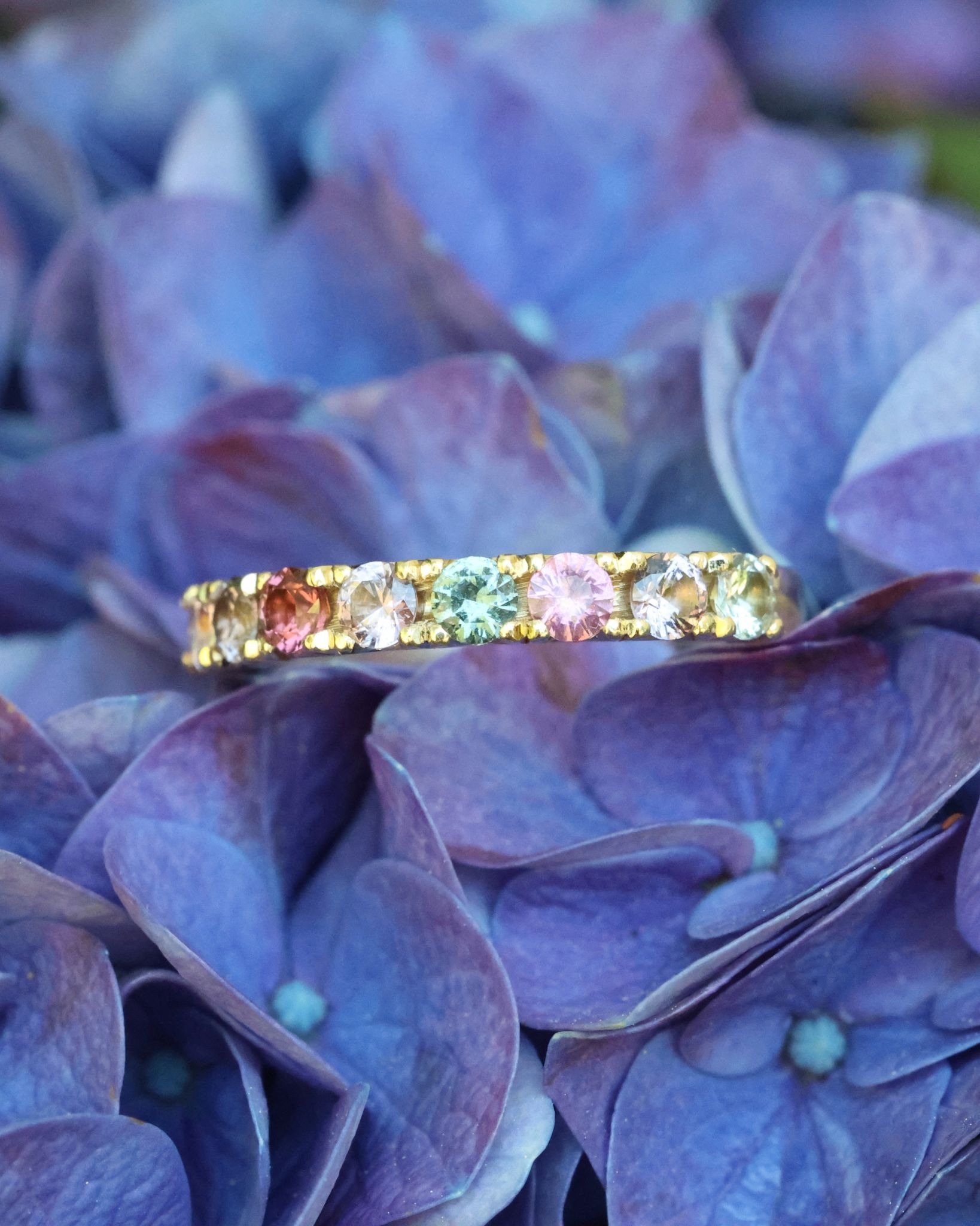 Gold gemstone eternity ring with multicolored stones resting on vibrant blue hydrangea petals.