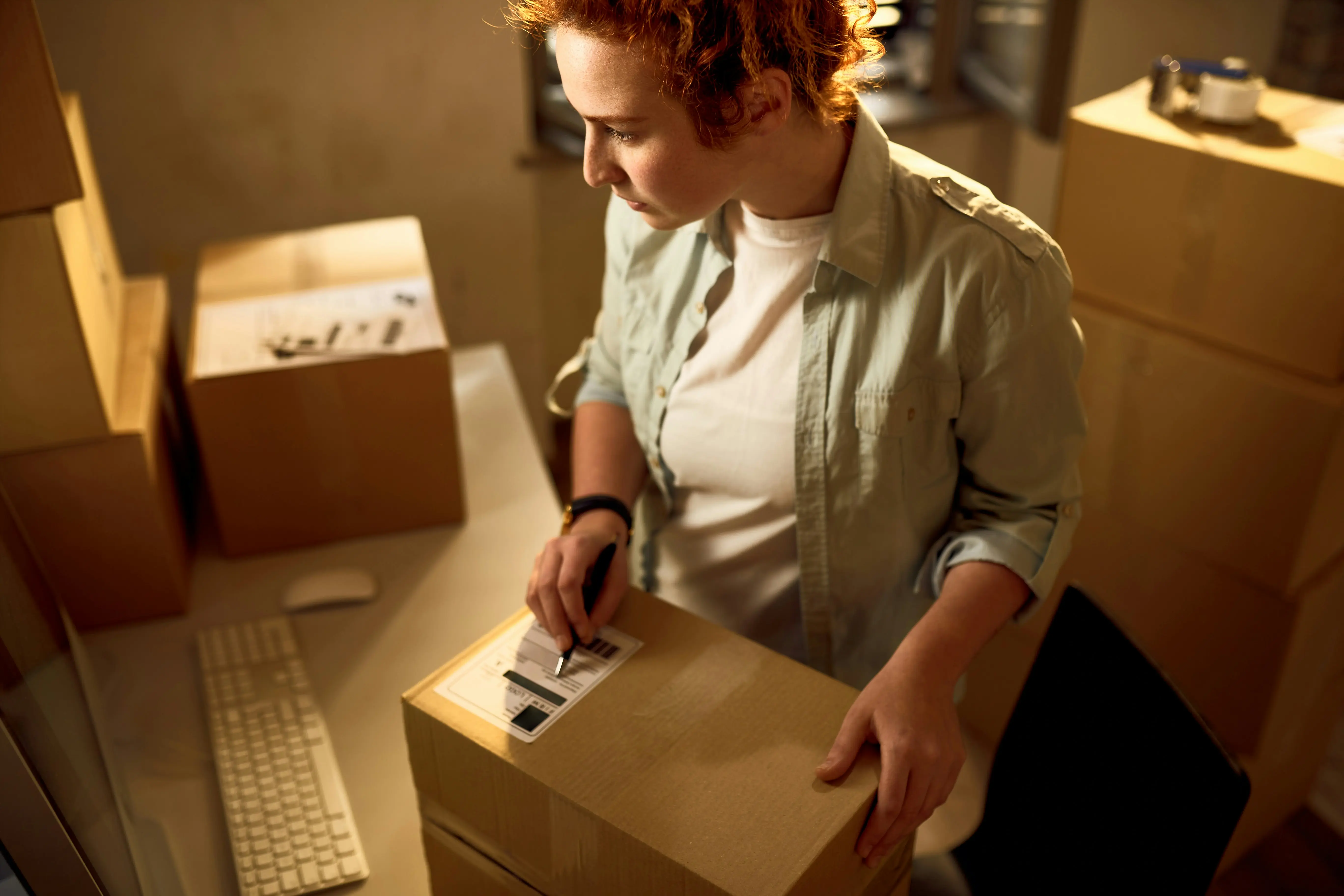 a woman with red hair looks at her computer while she has her hands on a carboard box in front of her
