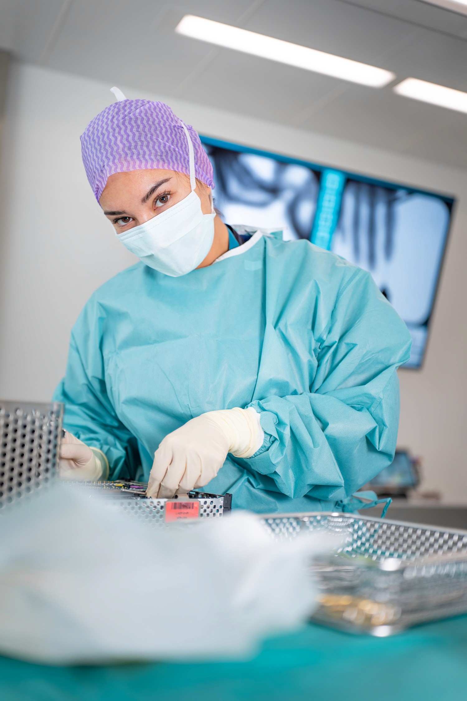 Eine Mitarbeiterin des Gesundheitswesens in blauem Kittel, lila Haarnetz und Gesichtsmaske sitzt im Operationssaal und bereitet Unterlagen vor, im Hintergrund ein Röntgenbild.