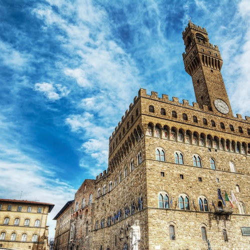 Storico edificio in pietra con finestre ad arco e un'alta torre dell'orologio sotto un cielo azzurro parzialmente nuvoloso. Statue adornano la facciata dell'edificio.