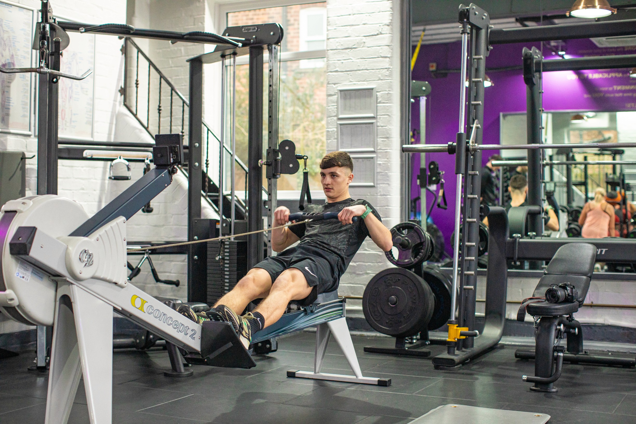 A man intensely using a rowing machine in a modern gym. Surroundings include weights and mirrors. The atmosphere is focused and energetic.