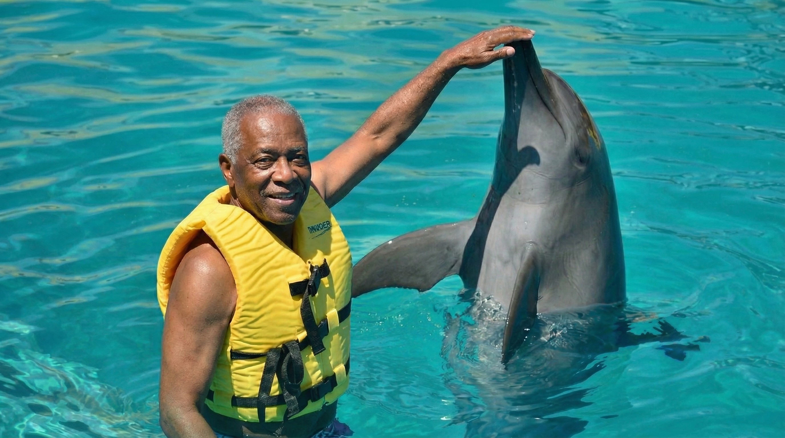 Smiling man in yellow life vest petting a dolphin.