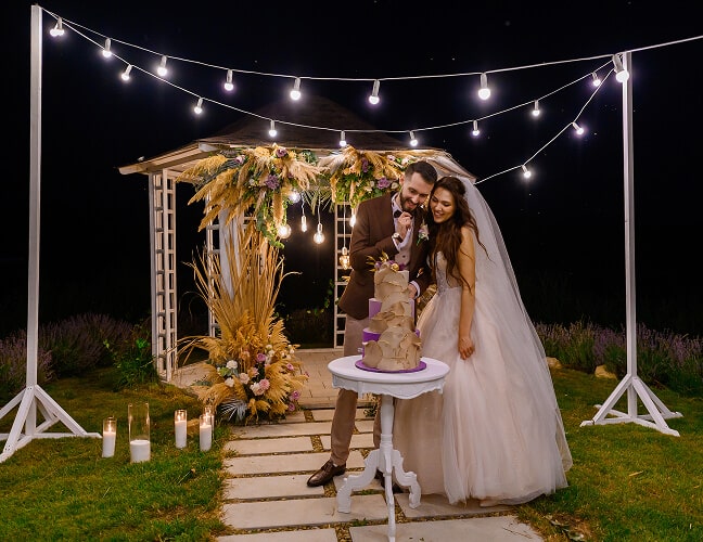 A bride in a white gown sits under twinkling lights, surrounded by candles and a floral arch in a nighttime setting.