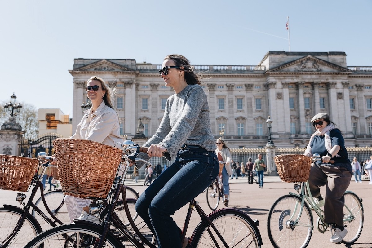 Smiling women riding vintage bicycles in front of Buckingham Palace on a Tally Ho Royal London Bike Tour 