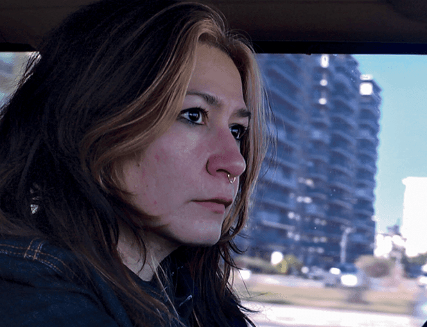 A woman with long brown hair and light-colored eyes is looking off to her right. She has a nose piercing and is wearing a dark jacket. A blurred cityscape with tall buildings is visible in the background, seen through a window. The woman’s expression is neutral or slightly pensive.