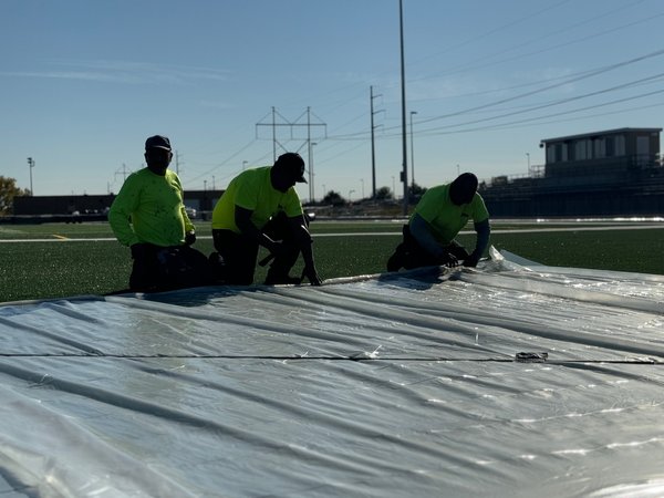 Three crew members in bright yellow shirts kneeling on plastic sheeting by a field