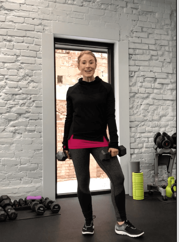 Heather Hausman in sportswear stands in a gym holding a dumbbell, surrounded by exercise equipment like kettlebells and a bright yellow water bottle, against a white brick wall.