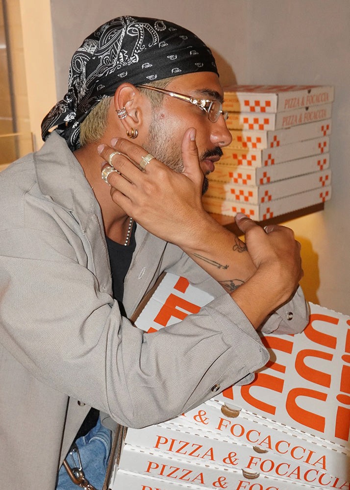 Person with a bandana and glasses holding a pizza box, with more pizza boxes stacked in the background. With silver rings.