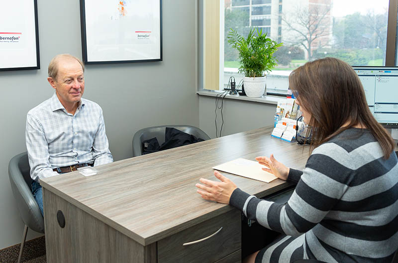 A man sits at a desk while a woman in stripes takes notes during a meeting in an office setting.