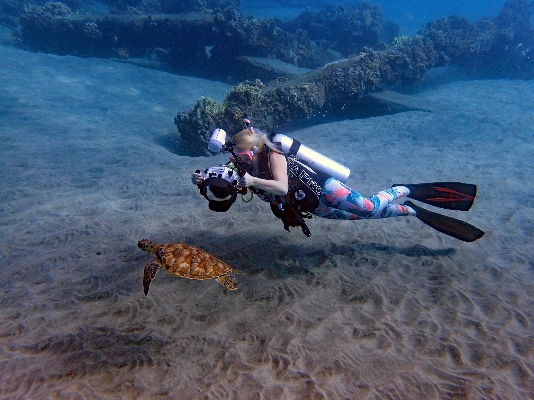 Scuba diver holding an underwater camera while swimming near a sea turtle above the sandy ocean floor.
