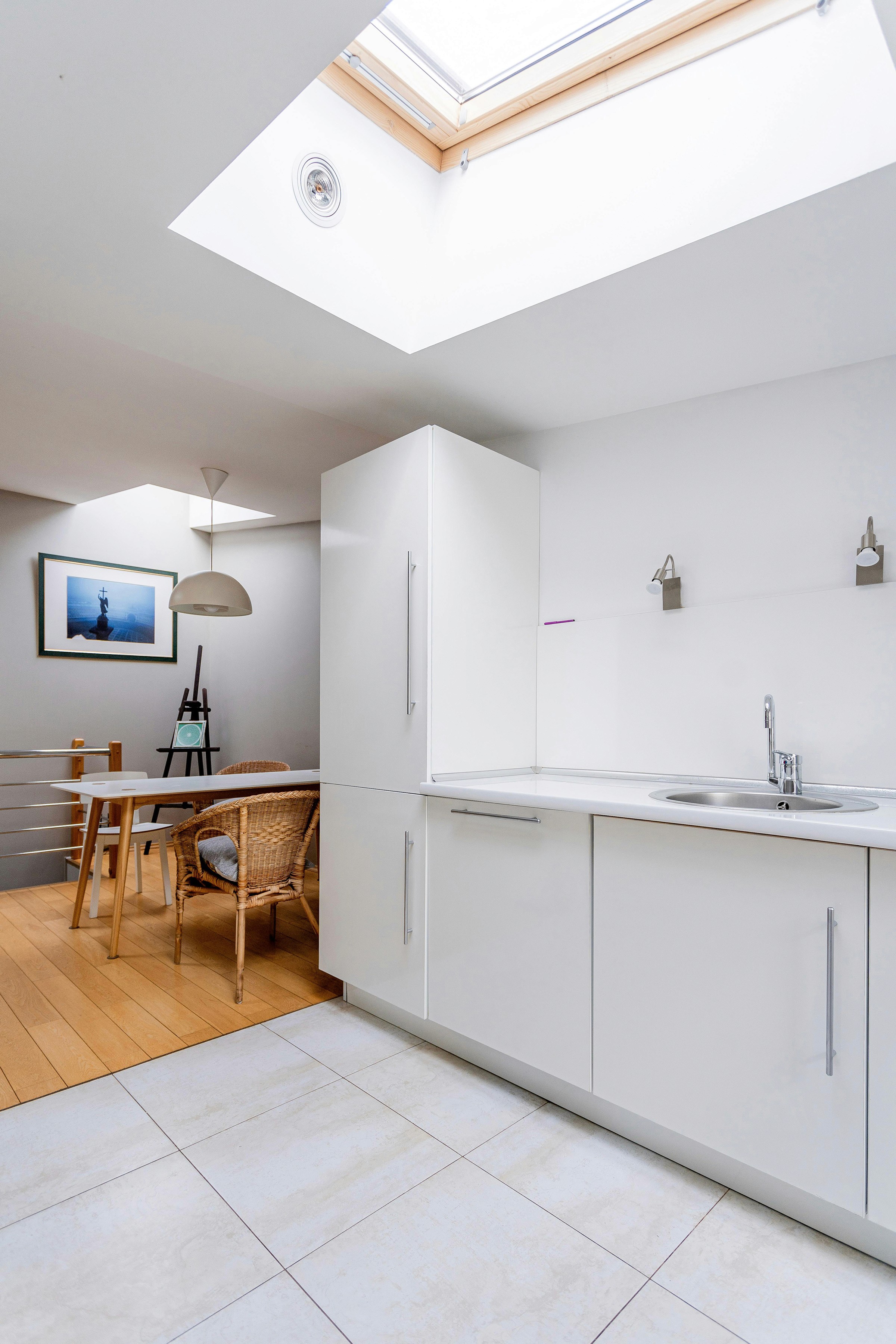 A modern kitchen with sleek white cabinets, a stainless steel sink, and a bright skylight illuminating the room. A cozy dining area with wicker chairs and minimalist decor is visible in the background