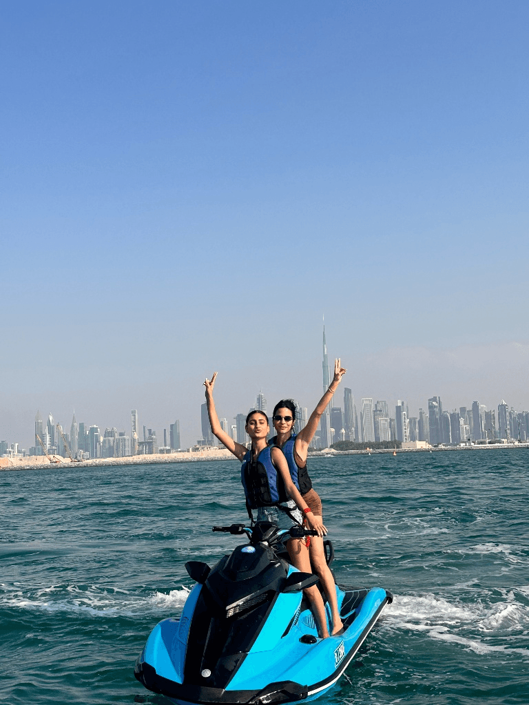 Two women posing with peace signs on a blue jet ski in the sea with the Dubai city skyline in the background.