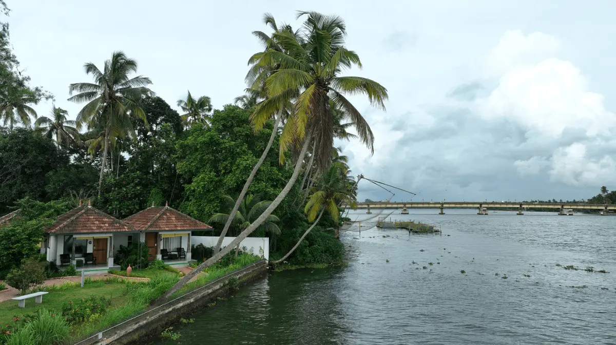 Lake View Villas at Vedic Village Kodungallur, twin terracotta-roofed cottages beside the lake with coconut palms and a traditional Chinese fishing net