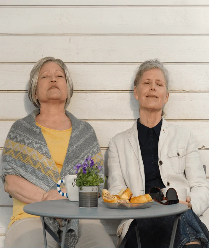 Två kvinnor sitter vid ett cafébord mot en vit husvägg och blundar mot solen