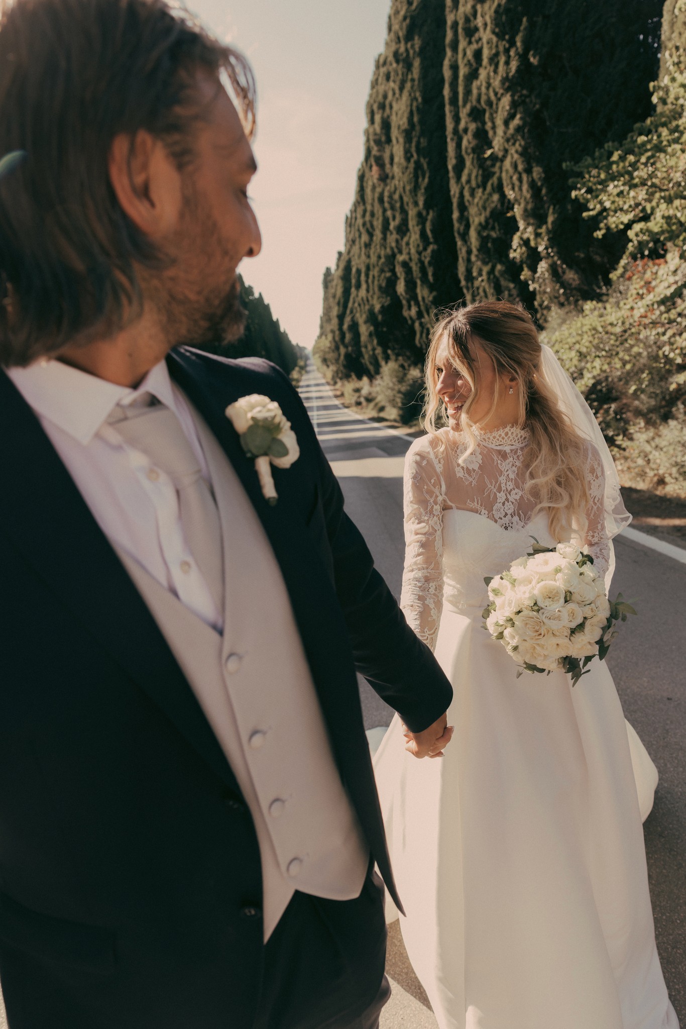 Sposi mano nella mano sul Viale dei Cipressi a Bolgheri, sposa in abito bianco con bouquet, sposo in tight