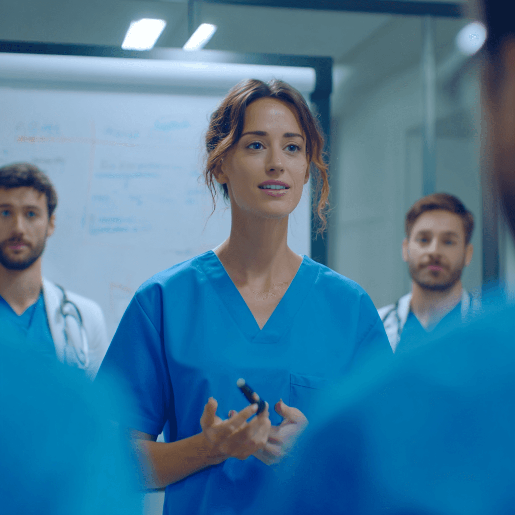 A female doctor in blue scrubs speaks confidently to a group, with two colleagues in the background. A whiteboard displays notes, suggesting a meeting.