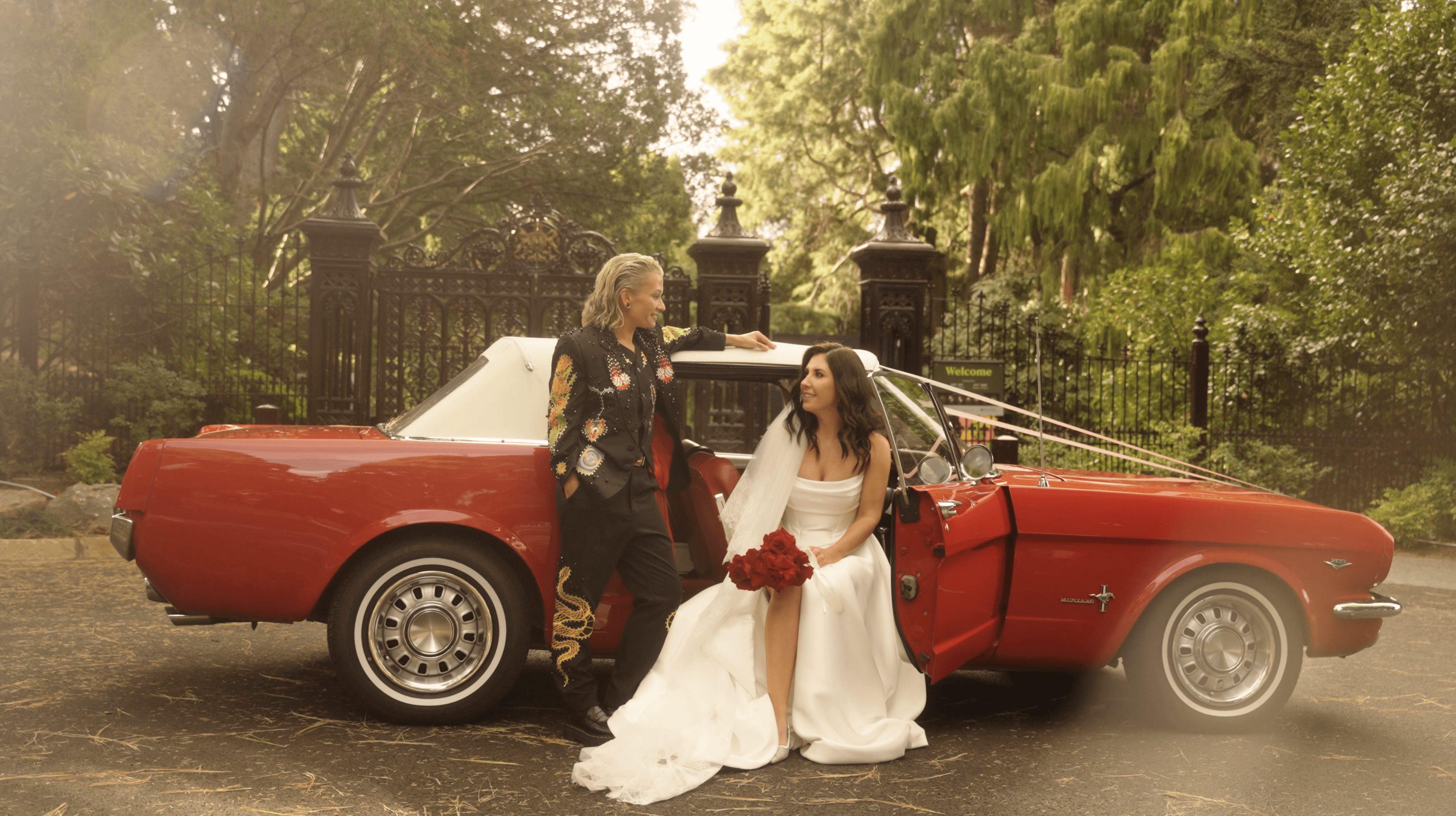 A joyful bride in a strapless white dress holds a bottle while celebrating with friends in a garden setting adorned with red roses and surrounded by a stone building.