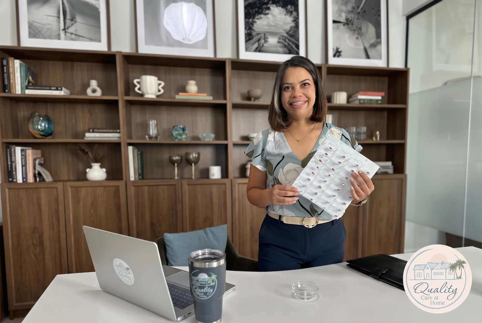 A woman stands at a white desk in a modern office in Bradenton, FL, holding a sheet with pills, with bookshelves in the background and a laptop open, promoting a health-conscious professional setting.