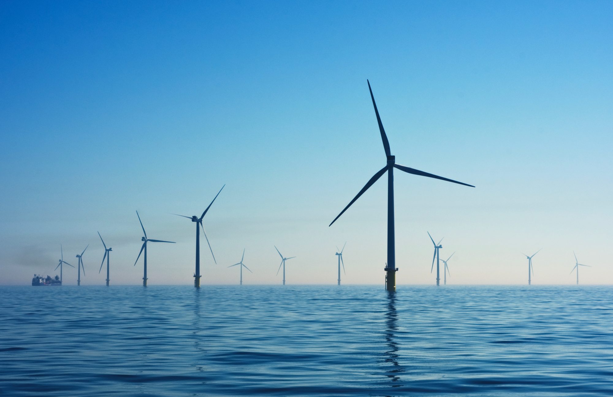 A wind farm with multiple turbines stands in the water against a clear blue sky.