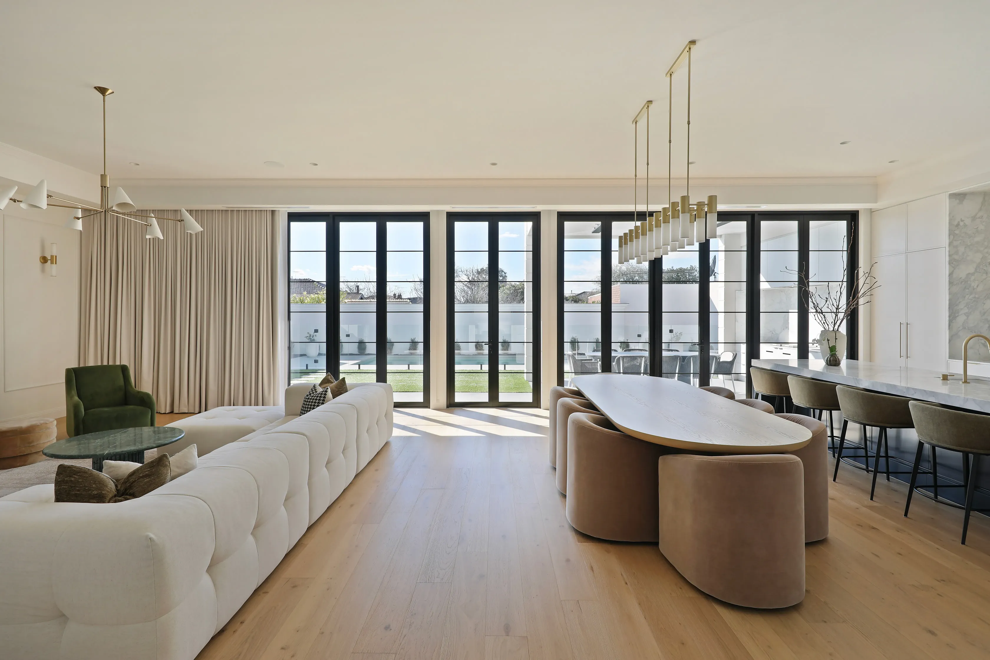 Open living and dining with a white tufted sofa, curved oval timber dining table with mauve velvet seating, brass linear pendant, oak floors and a wall of black-framed doors to the pool terrace