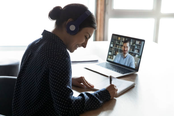 Giovane donna con cuffie che sorride durante una lezione online, prendendo appunti su un quaderno mentre parla con il docente collegato in videochiamata sul laptop.