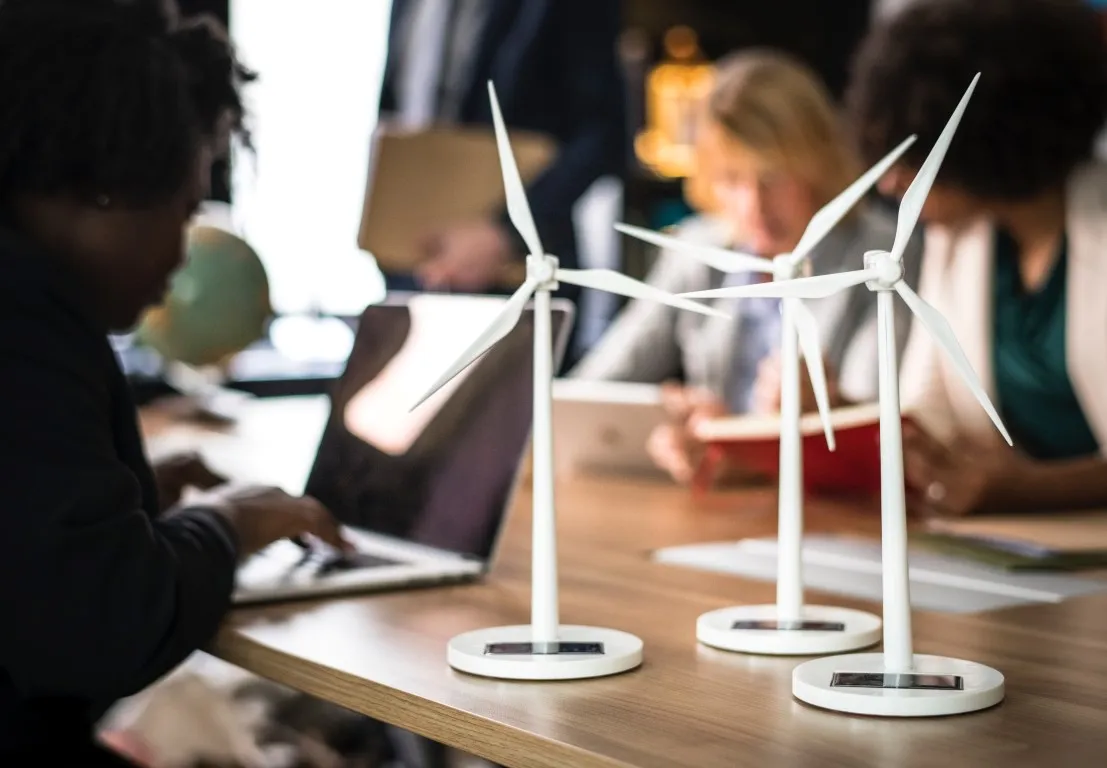 Three small white model wind turbines are displayed on a wooden table in the foreground, with blurred figures of people working on laptops in the background, suggesting discussions around renewable energy, sustainable technology, or eco-friendly business.
