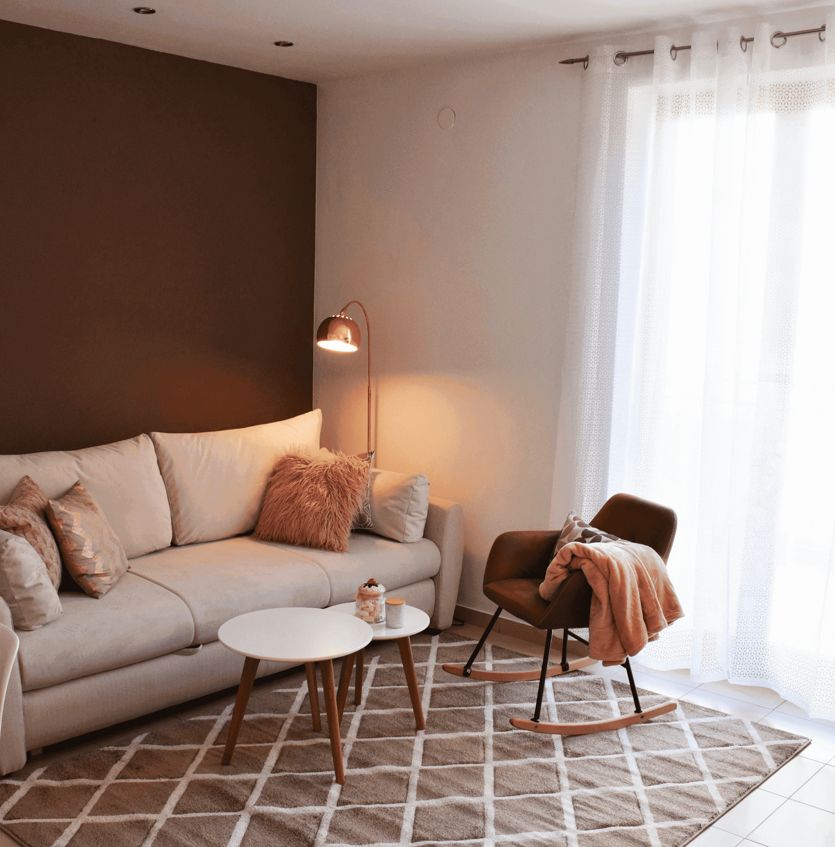 A cozy living room featuring a beige sofa with plush pillows and a furry throw, a modern rocking chair draped with a soft pink blanket, illuminated by warm light from a stylish floor lamp, set against a contrast of dark brown and white walls, with sheer curtains letting in natural light over a geometric patterned area rug.