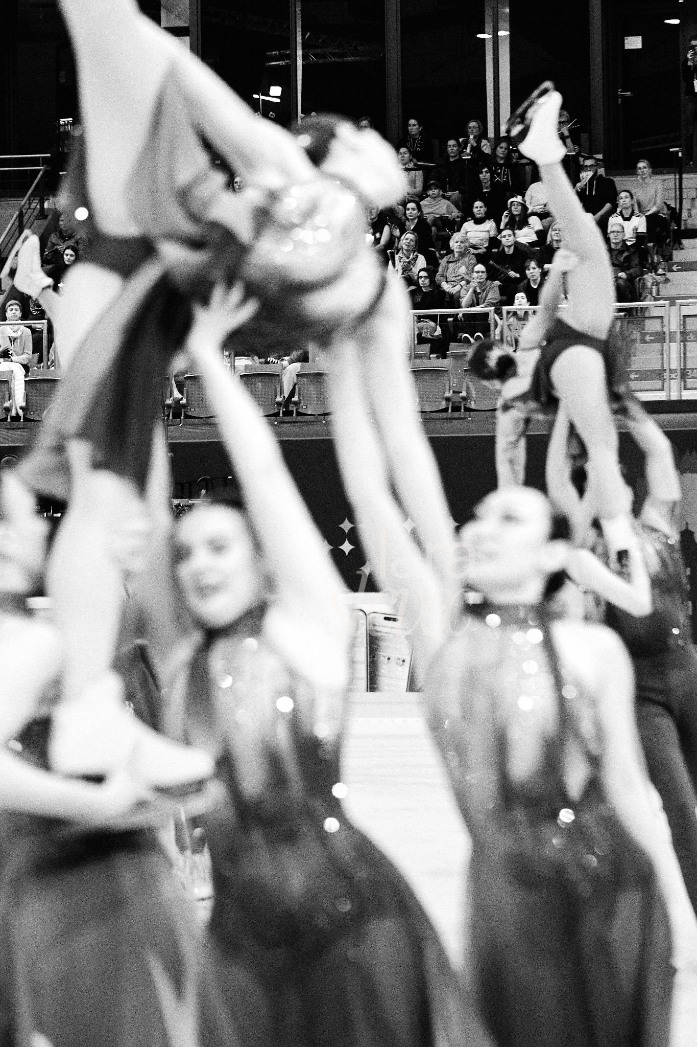 Schwarz-Weiß Bild im Analog Stil. Zwei Hebungen im Synchroneiskunstlauf in Unschärfe im Vordergrund. Im Hintergrund sitzt die staunende Tribüne. Team Nova. ISU Synchronized Skating Championships 2026 in Salzburg. Fotografiert von Lara Slevec.