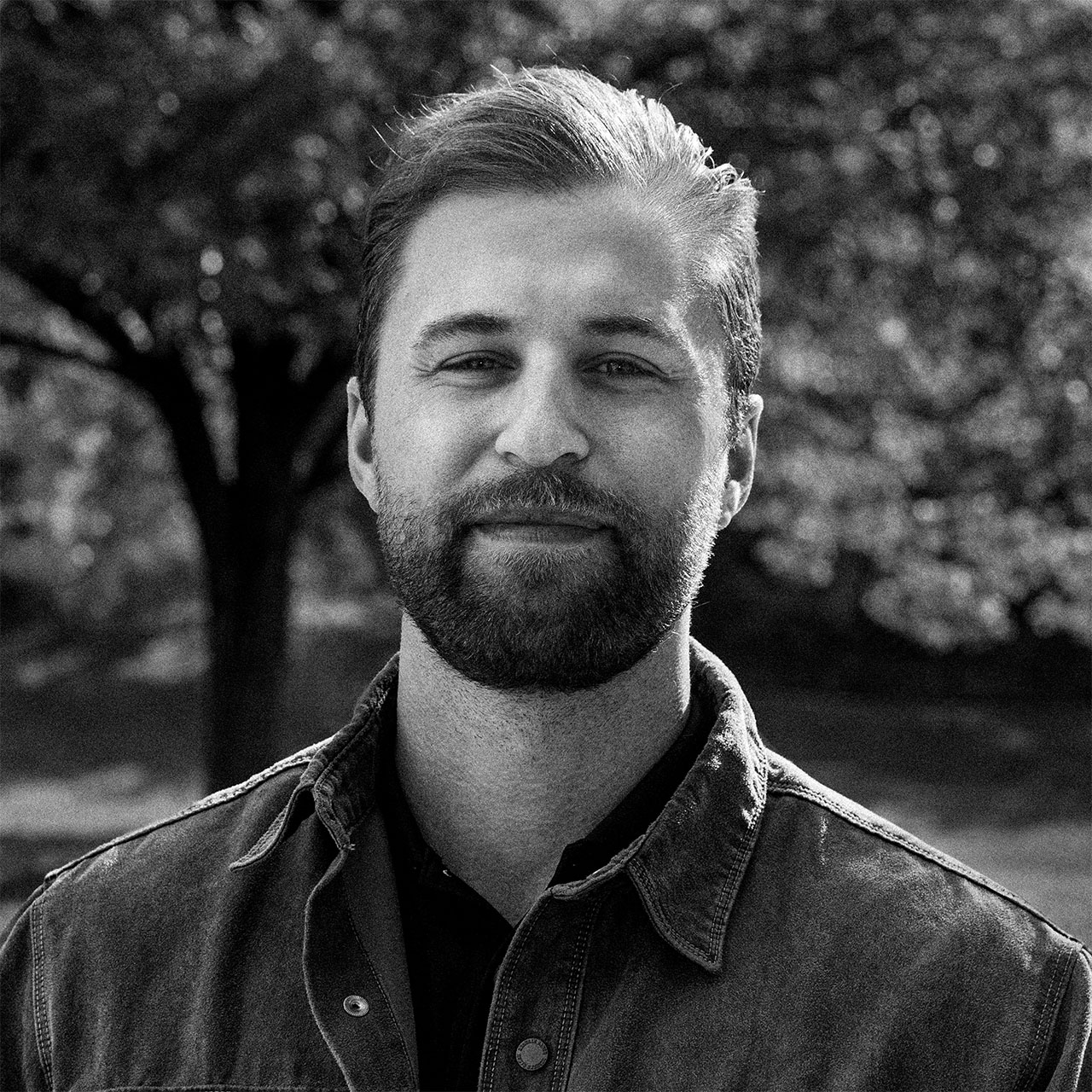 An architect standing at a desk in a modern studio, reviewing architectural models and documents. Shelves filled with books and materials line the background, with natural light coming through large windows.