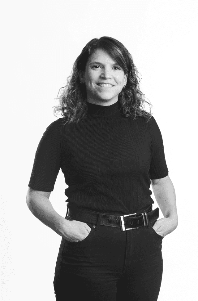 Black and white portrait of a smiling woman with curly hair, wearing a black turtleneck and pants, hands in pockets, against a white background.