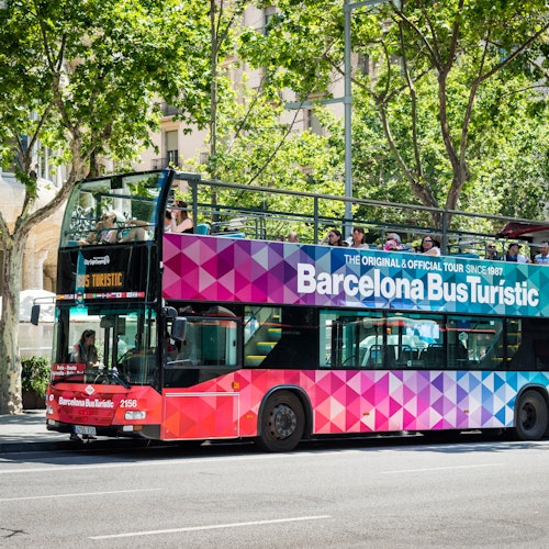 Un autobús de dos pisos de colors vius etiquetat com "Barcelona Bus Turístic" amb passatgers a la part superior, estacionat en un carrer envoltat d'arbres.