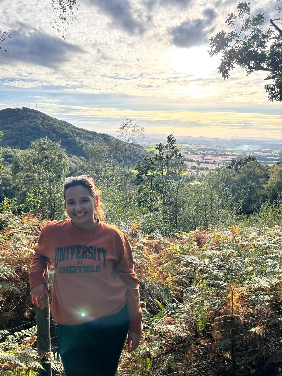 Una persona con una camiseta negra y pantalones de color claro está de pie en la cima rocosa de una montaña al atardecer, sosteniendo una cámara frente a su cara para tomar una foto. Un mar de nubes se extiende detrás de ellos, iluminado por la luz dorada del sol poniente.