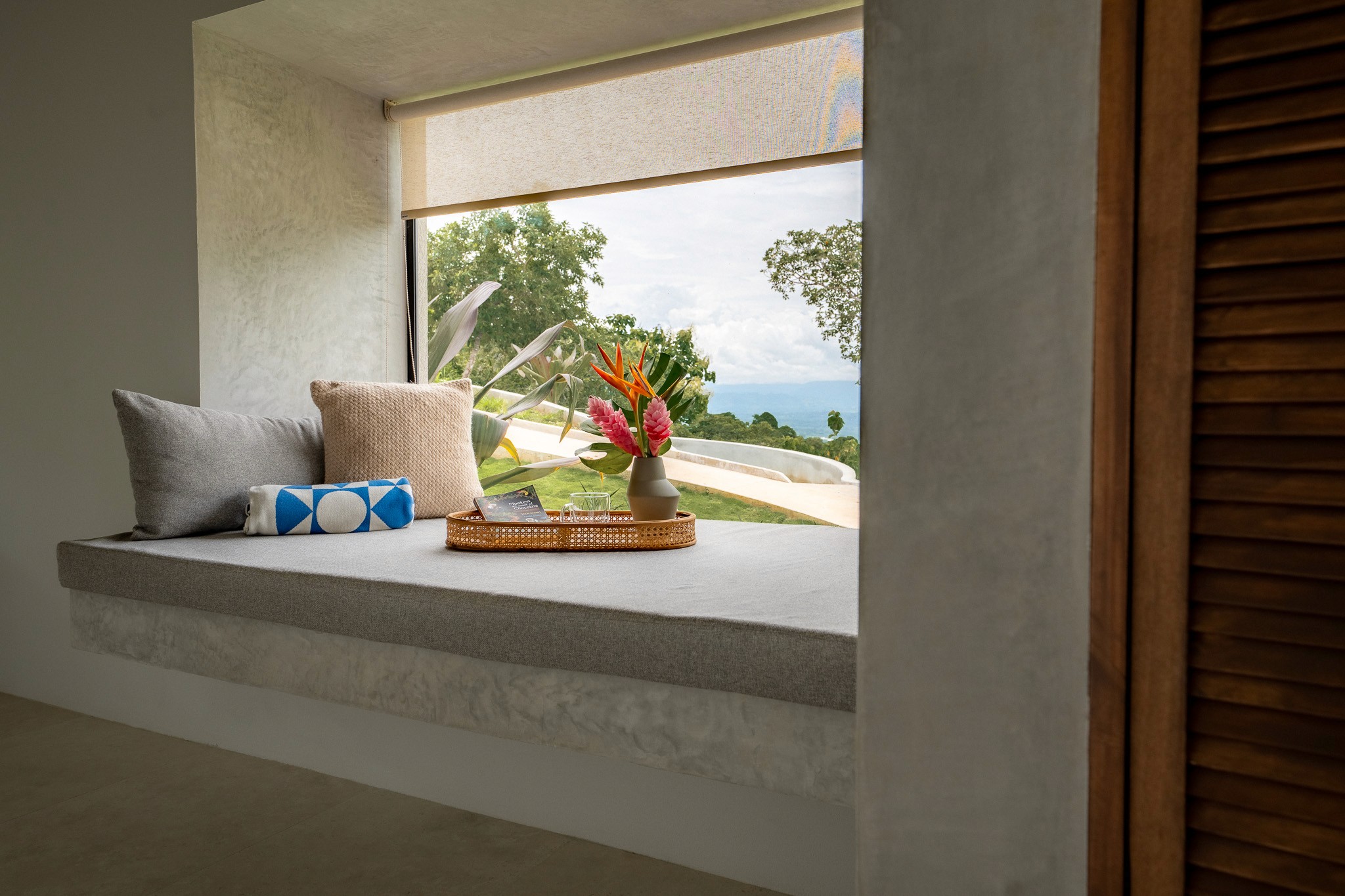 Cozy built-in window seat nook with pillows, a book, and tropical flowers overlooking the lush landscape and distant ocean.