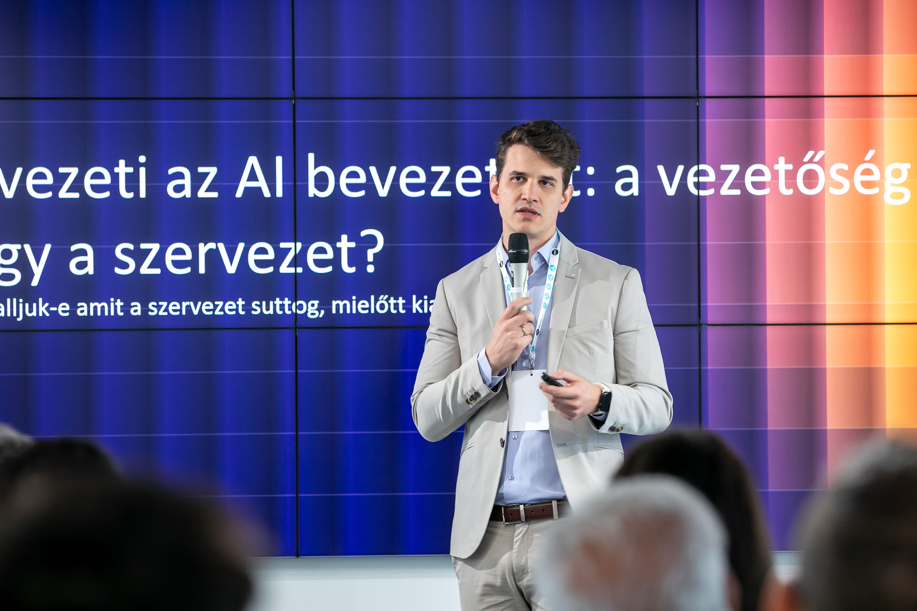 Irodai tárgyalás elmosódott háttérrel – az előtérben toll, szemüveg és grafikonokat tartalmazó dokumentumok láthatók, a háttérben laptopok előtt ülő és álló üzleti szakemberek tárgyalnak