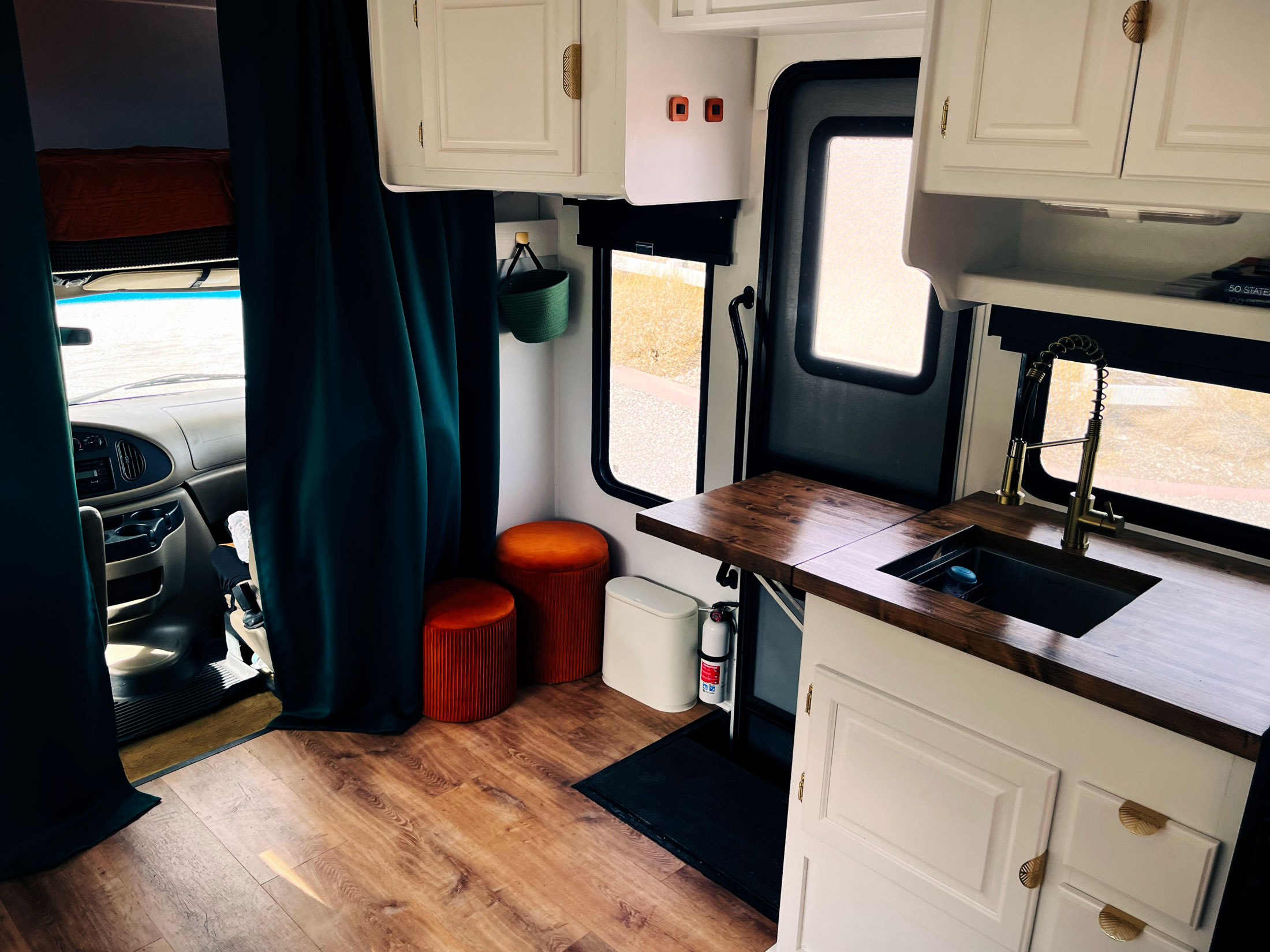 RV kitchen interior with wood flooring, white cabinets, and a dark wood worktop. Features dark curtains, orange stools, and a fire extinguisher. Cozy atmosphere.