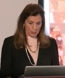 A photo of a woman wearing a suit and standing at a laptop while giving a lecture.