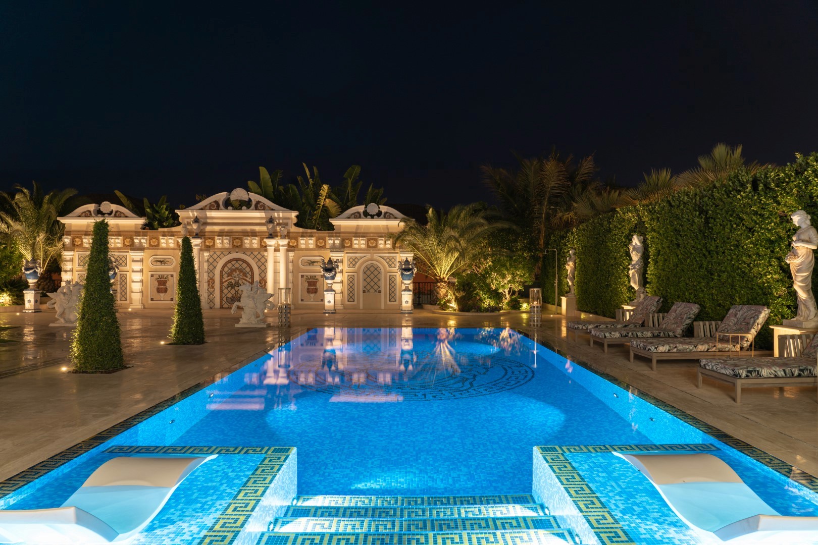 Luxurious nighttime view of an ornate outdoor swimming pool, surrounded by elegant statues, lush greenery, and classical architecture, illuminated by warm lighting.
