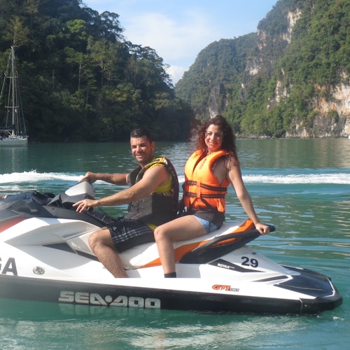 一男一女坐在水上摩托艇上，背景是一個風景如畫的海灣，有懸崖和綠樹。他們都笑著，穿著救生衣。