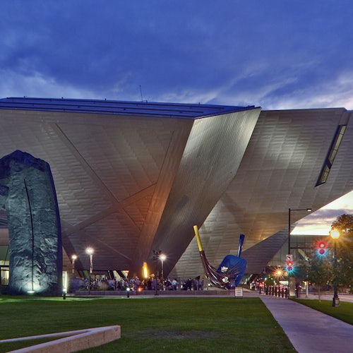 Modern angular building with unique architecture, illuminated at dusk. People are gathered near its entrance and sculptures.