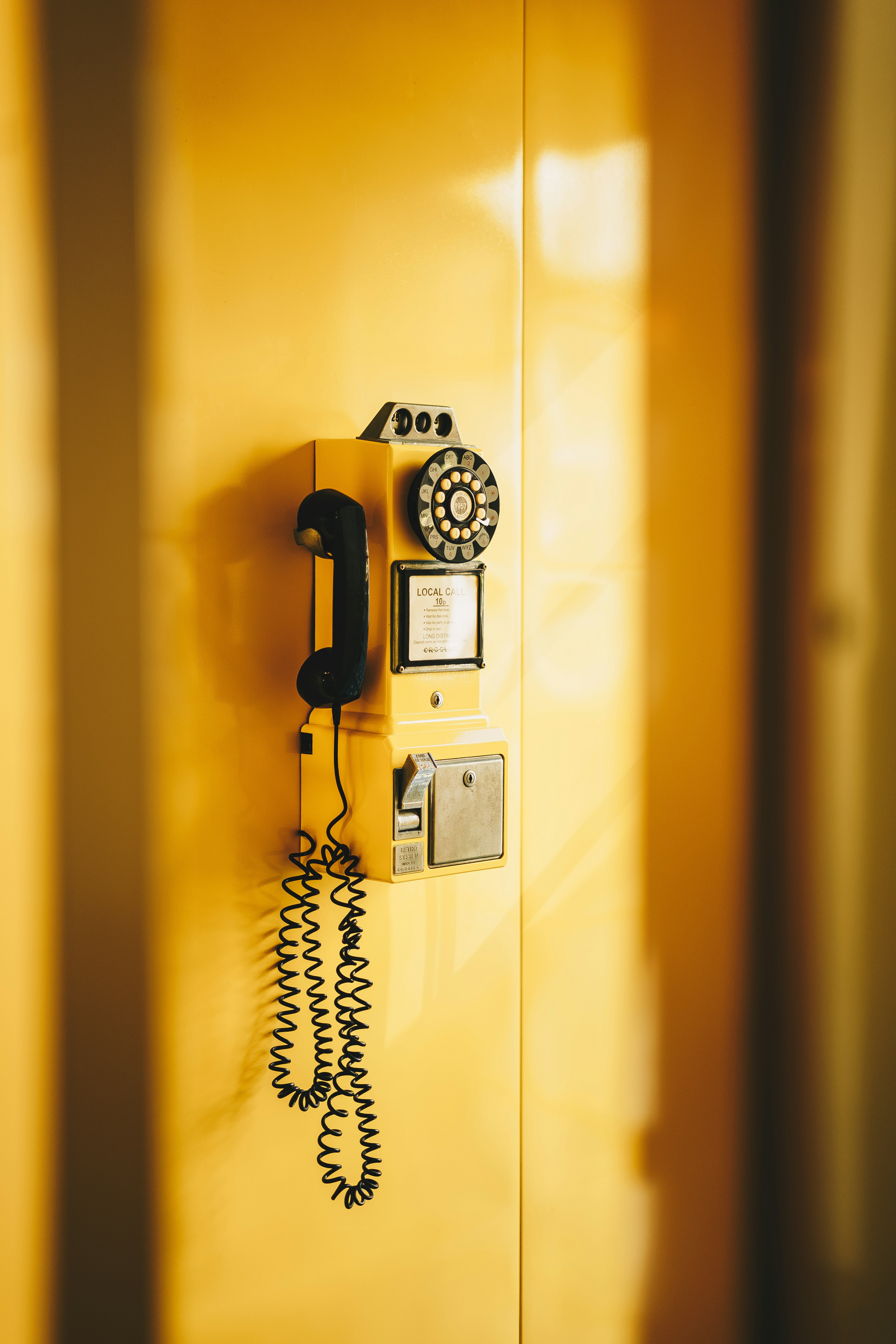 A vintage yellow rotary payphone mounted on a matching yellow wall, with soft sunlight casting shadows across the scene.