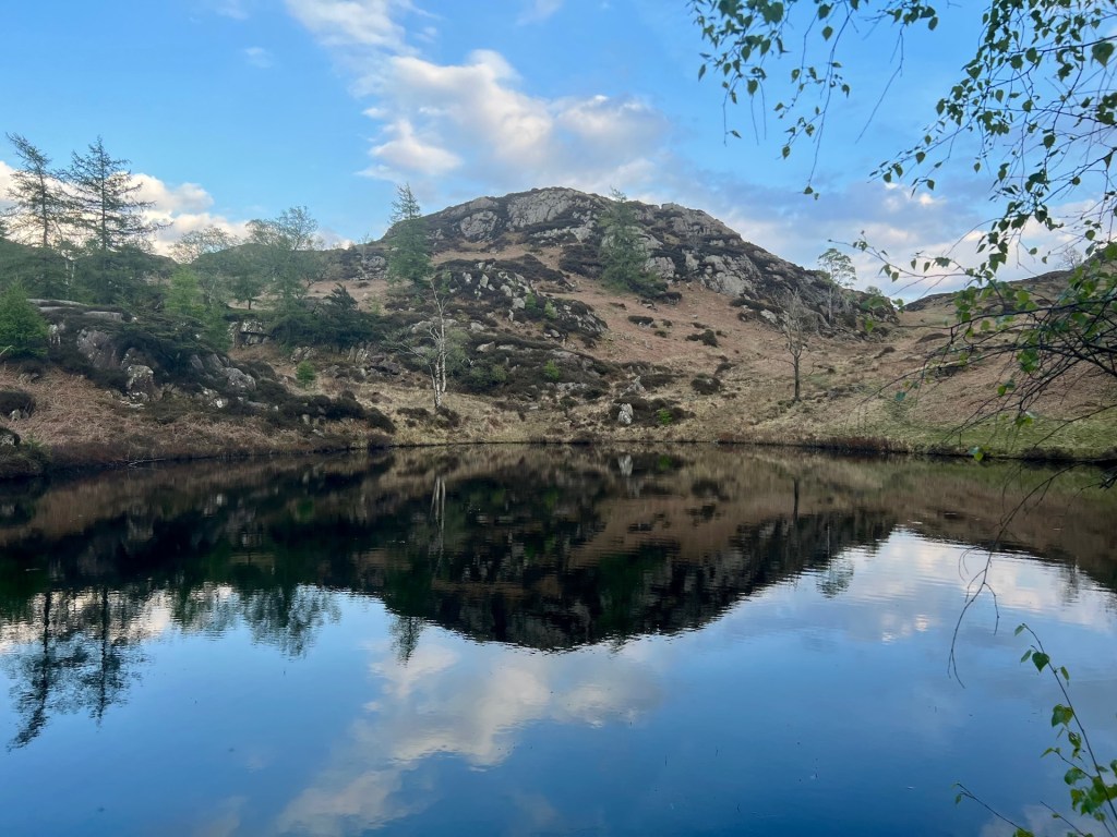 The Holme Fell reservoir, still as a mirror, reflecting the sky above and the hills behind.