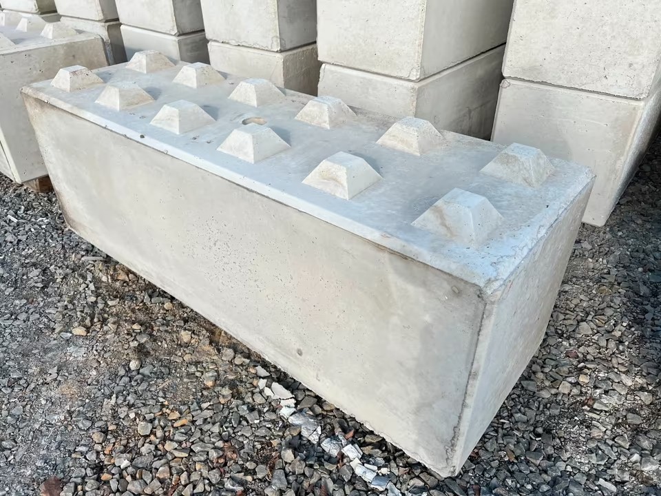 Grand bloc rectangulaire en béton blanc avec plots pyramidaux sur le dessus, similaire à une brique Lego géante, stocké sur un sol de graviers avec d'autres blocs identiques en arrière-plan