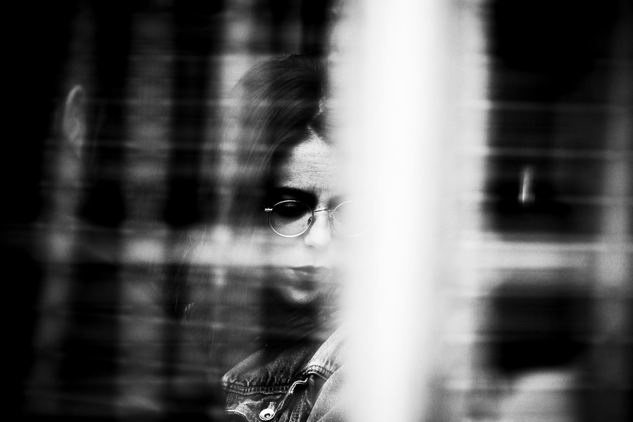 Woman seen through reflections and glass wearing round glasses, black and white street photography portrait.