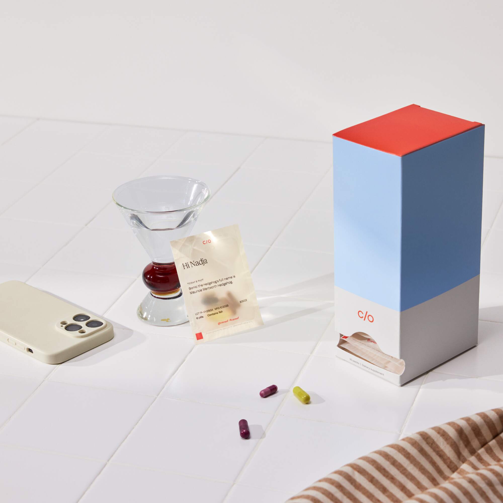 Care/of product display with a personalized vitamin pack, smartphone, glass of water, and supplement box on a white surface.