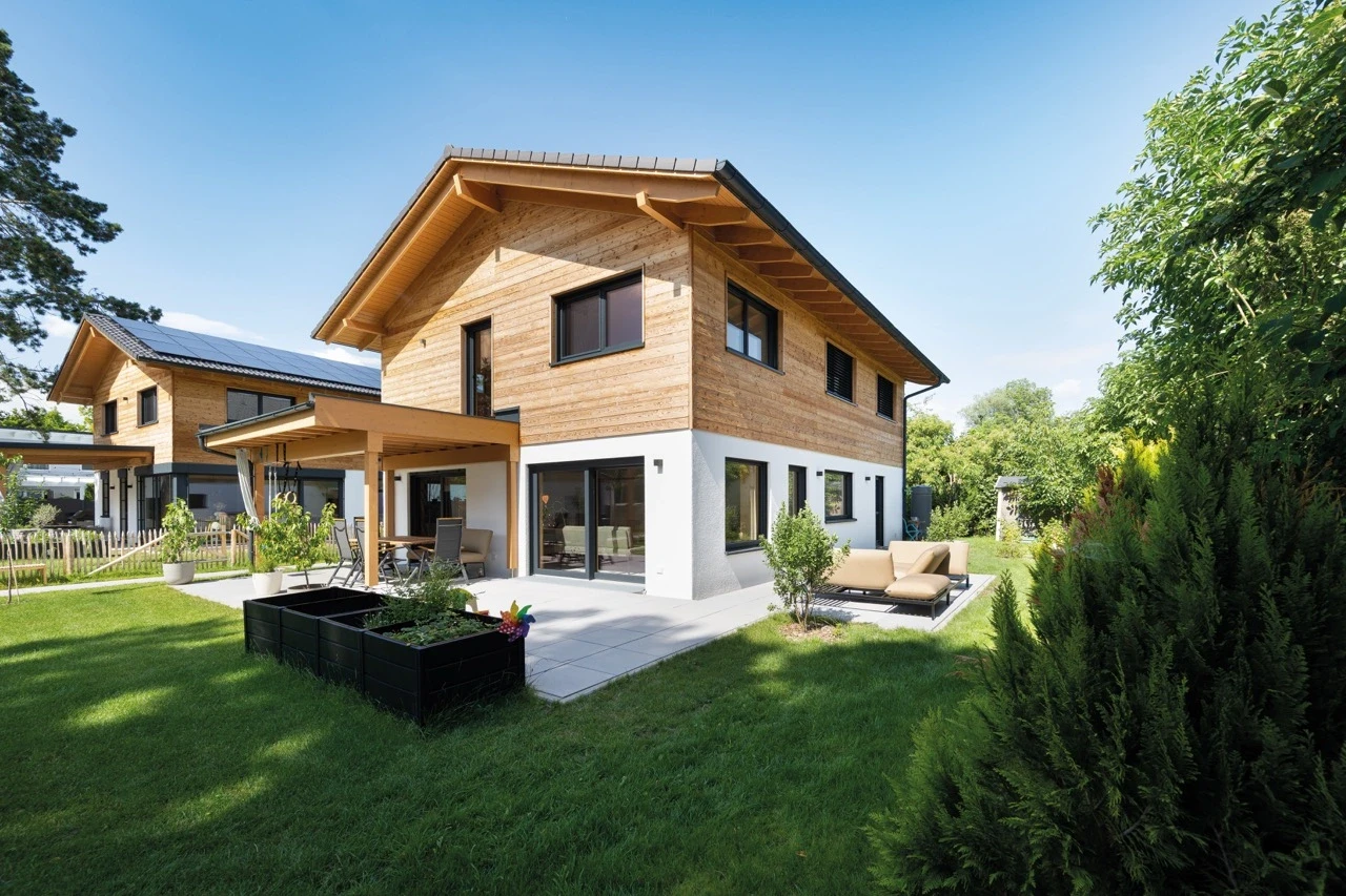 Modern wooden house with patio, surrounded by lush greenery.