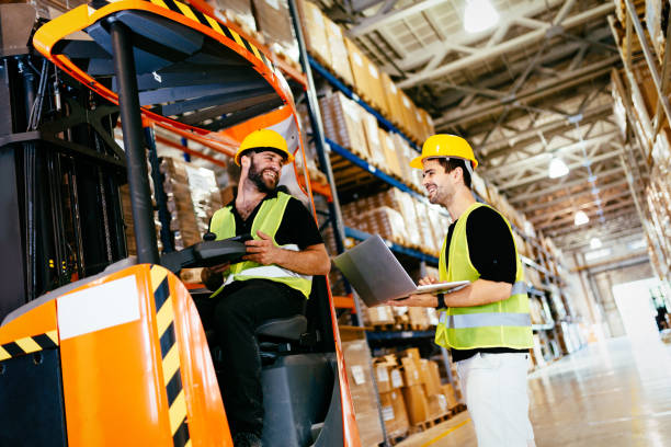 two people wearing hard hats and hugh vis vest in a warehouse and one is driving a forklift