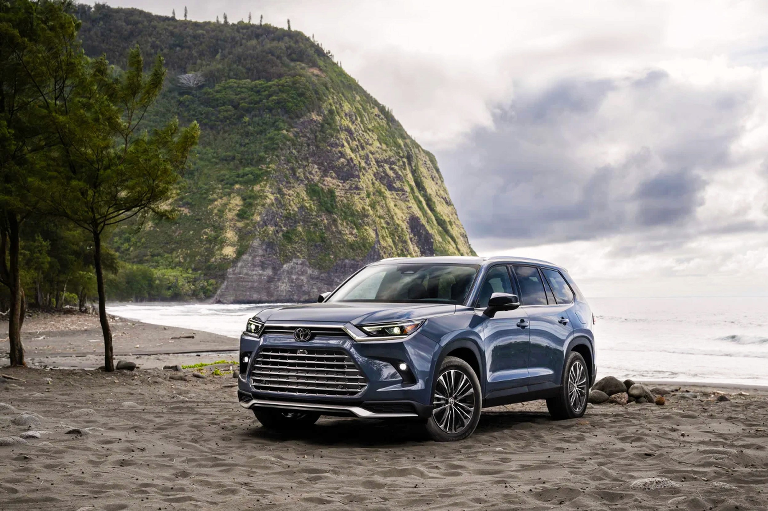 A 2025 Toyota Grand Highlander is parked on a beach. Weather is slightly overcase and there is a mountain in the background.