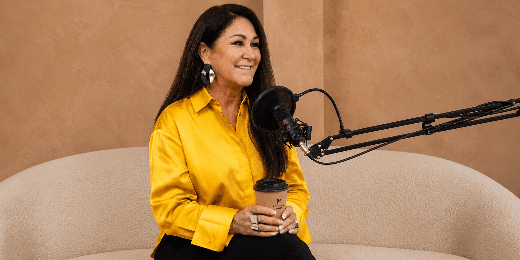 Traycee Mayer, a woman on a couch, holds a microphone and coffee, symbolizing her focus on connection in leadership.