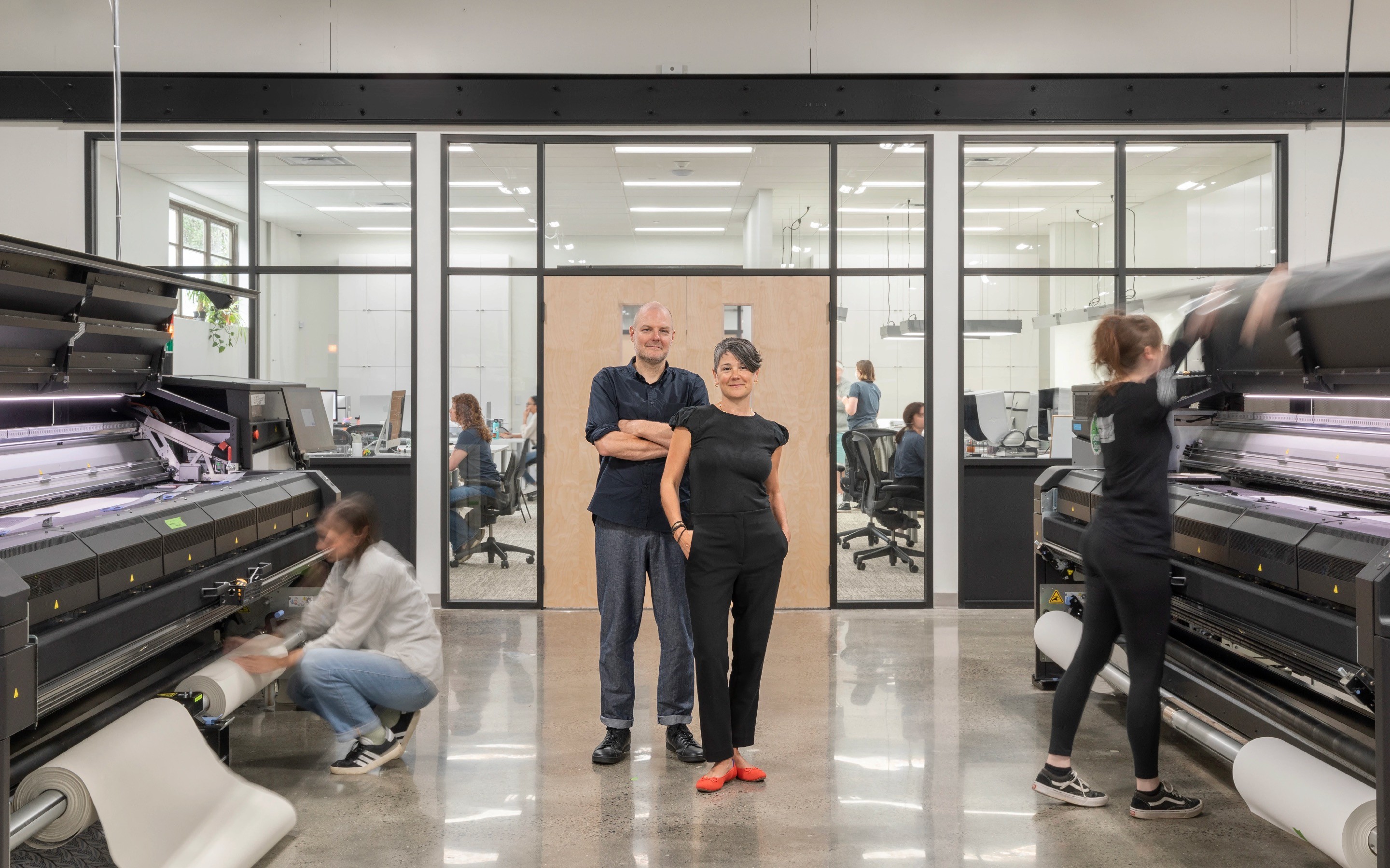 Kyra and Rob Hartnett, owners of twenty two wallpaper, standing in front of large wallpaper printers.
